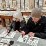 Tracey Gemmell using traditional Chinese paint brushes to copy symbols with Chinese art on wall behind.