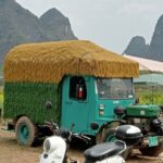 A green truck with a thatched roof on the banks of the Yulong River, mountains in the background.